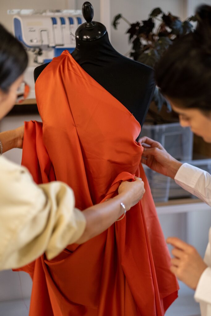 Journal Two designers collaborate in a fashion studio, adjusting fabric on a mannequin.